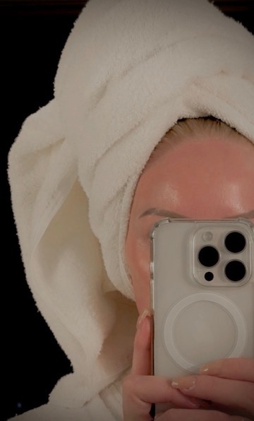 Soft, glowing post-treatment skincare photo of a woman with a towel wrapped around her hair, holding a phone in front of her face with radiant, dewy skin visible.